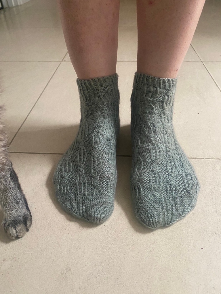 White woman’s ankles and feet in daintily cabled ankle socks. A cat’s toes pose alongside. 