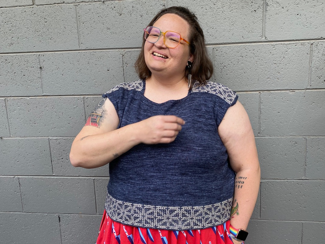 A white woman with short brown hair, tattoos, and glasses, modeling the Boxy Superlite Top in front of a gray concrete block wall
